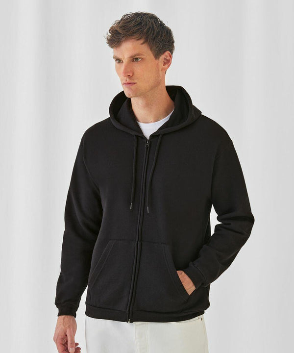 A person with short hair is wearing a black B&C Collection BA412 hoodie over a white shirt. The B&C Collection BA412 hoodie has a zipper down the front and drawstrings. The individual has their left hand in the B&C Collection BA412 hoodie pocket and is standing against a white background.