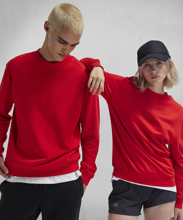 Two people stand before a plain light grey background, both in bright red B&C ID.332 crew sweatshirts by B&C Collection. The left person has short blonde hair; the right wears a black cap, has fair hair, and black shorts.