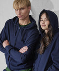 Two people stand in front of a plain light wall wearing B&C Collection's B&C ID.334 zip hoodies—one with arms crossed and a neutral look, the other with long hair, a slight smile, and the hood up. Both hoodies are navy blue.