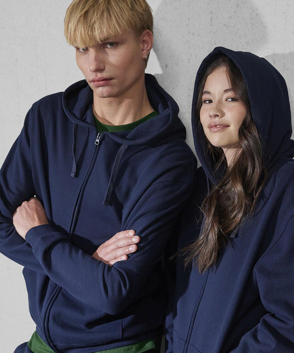 Two people stand in front of a plain light wall wearing B&C Collection's B&C ID.334 zip hoodies—one with arms crossed and a neutral look, the other with long hair, a slight smile, and the hood up. Both hoodies are navy blue.