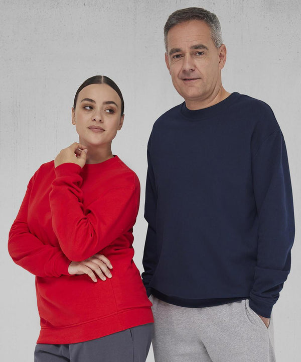 A woman in a red B&C Collection B&C ID.222 jumper and grey trousers stands next to a man in a navy B&C ID.222 jumper and light grey trousers, both facing forward with neutral expressions against a light grey textured background.