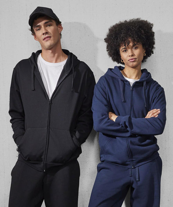 A man in a black B&C Collection B&C ID.224 zip hoodie and a woman in a navy blue one stand side by side against a light grey wall, both looking at the camera with neutral expressions.