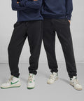 Two people stand side by side in navy sweatshirts, B&C Collection B&C ID.000 joggers, and white trainers with green accents against a plain light grey background. Their faces are not visible.