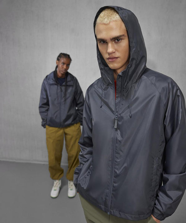 Two people stand indoors against a grey wall, both wearing B&C Collection's Reset lined windcheaters. The person in front has blond hair; the one behind has darker hair, khaki trousers, and white shoes.