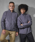 Two people stand side by side against a grey concrete wall, both wearing matching B&C Collection B&C Reset polar fleece zip-up jackets—one with short hair and beige trousers, the other with curly hair and black trousers.