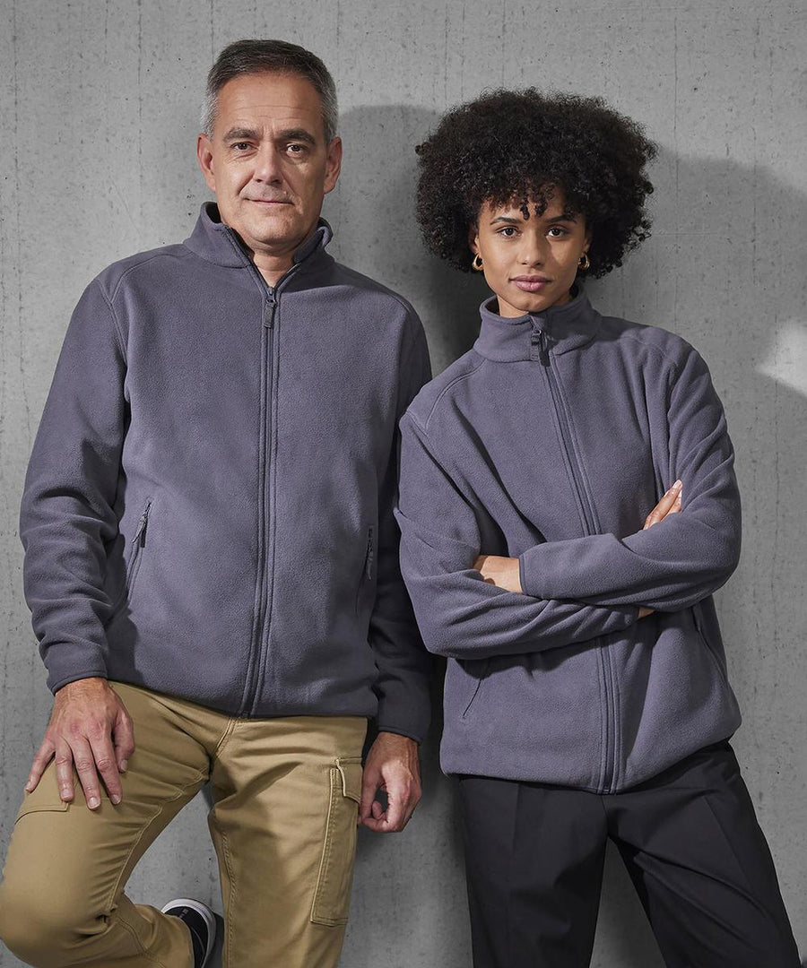 Two people stand side by side against a grey concrete wall, both wearing matching B&C Collection B&C Reset polar fleece zip-up jackets—one with short hair and beige trousers, the other with curly hair and black trousers.