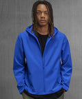 A person with long braided hair stands against a grey concrete-textured background, wearing a bright blue B&C Collection B&C Reset 3Lr hooded softshell jacket over a black shirt with their hands in their pockets.