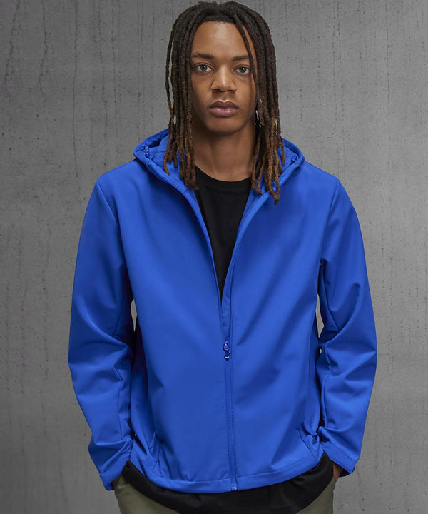 A person with long braided hair stands against a grey concrete-textured background, wearing a bright blue B&C Collection B&C Reset 3Lr hooded softshell jacket over a black shirt with their hands in their pockets.