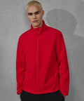 A person with short blond hair wears a bright red B&C Collection B&C Reset 3Lr softshell jacket, standing in front of a grey concrete wall and looking at the camera with a neutral expression.