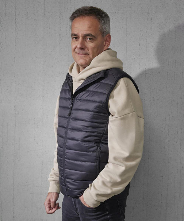 A middle-aged man with short grey hair stands against a plain grey wall, wearing a beige hoodie and the B&C Collection B&C Reset bodywarmer, looking at the camera with a neutral expression.