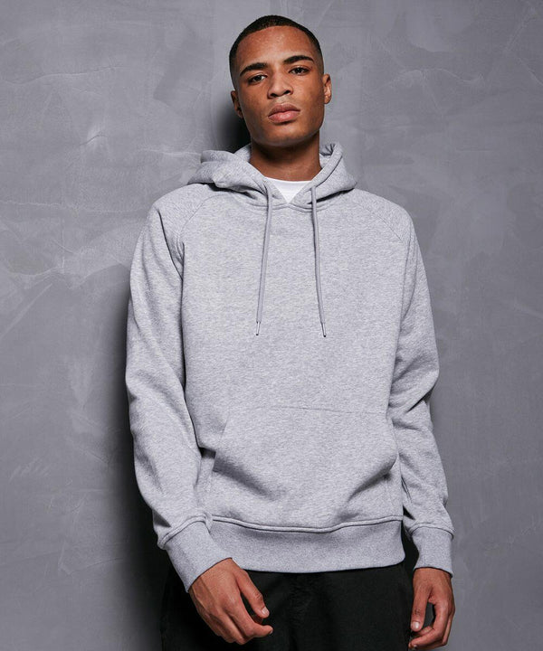 A person stands against a textured gray wall, wearing a Build Your Brand Basic BB001 light gray hoodie with the hood down and drawstrings hanging loose. They have short hair and a neutral facial expression, with their left arm slightly bent and right arm resting at their side.