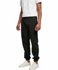 A person wearing a white T-shirt, BB002 black jogger pants from Build Your Brand Basic, and white sneakers stands against a plain white background. The BB002 black jogger pants from Build Your Brand Basic have an elastic waistband and cuffs at the ankles.
