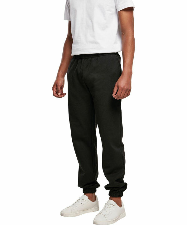 A person wearing a white T-shirt, BB002 black jogger pants from Build Your Brand Basic, and white sneakers stands against a plain white background. The BB002 black jogger pants from Build Your Brand Basic have an elastic waistband and cuffs at the ankles.