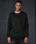 A person with a serious expression stands against a dark, textured background wearing a black BB003 long-sleeved shirt by Build Your Brand Basic and dark jeans. They are looking directly at the camera with their arms relaxed at their sides.