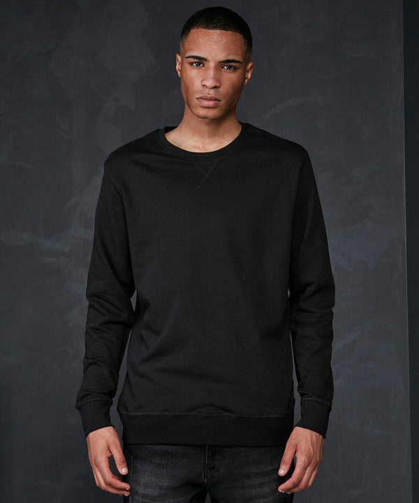 A person with a serious expression stands against a dark, textured background wearing a black BB003 long-sleeved shirt by Build Your Brand Basic and dark jeans. They are looking directly at the camera with their arms relaxed at their sides.