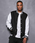 A person stands against a textured grey background, wearing a black varsity jacket with white sleeves and cuffs, a white shirt underneath, and black pants. They have a neutral expression and look directly at the camera, showcasing the BB004 from Build Your Brand Basic.
