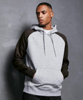 A person wearing a light gray **BB005 by Build Your Brand Basic** with dark gray sleeves stands against a textured, dark gray wall. The person has their hands in the hoodie pocket and looks off to the side with a neutral expression.