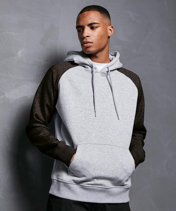 A person wearing a light gray **BB005 by Build Your Brand Basic** with dark gray sleeves stands against a textured, dark gray wall. The person has their hands in the hoodie pocket and looks off to the side with a neutral expression.