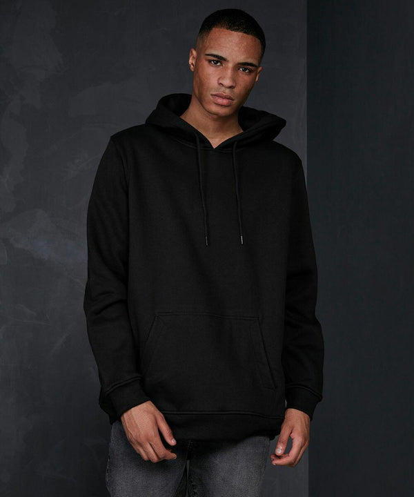 A person stands against a dark gray backdrop wearing a black Build Your Brand Basic BB006 hoodie with the hood down and hands in the front pocket. They have a neutral expression and are dressed in dark gray jeans. The overall atmosphere is minimalist and casual.