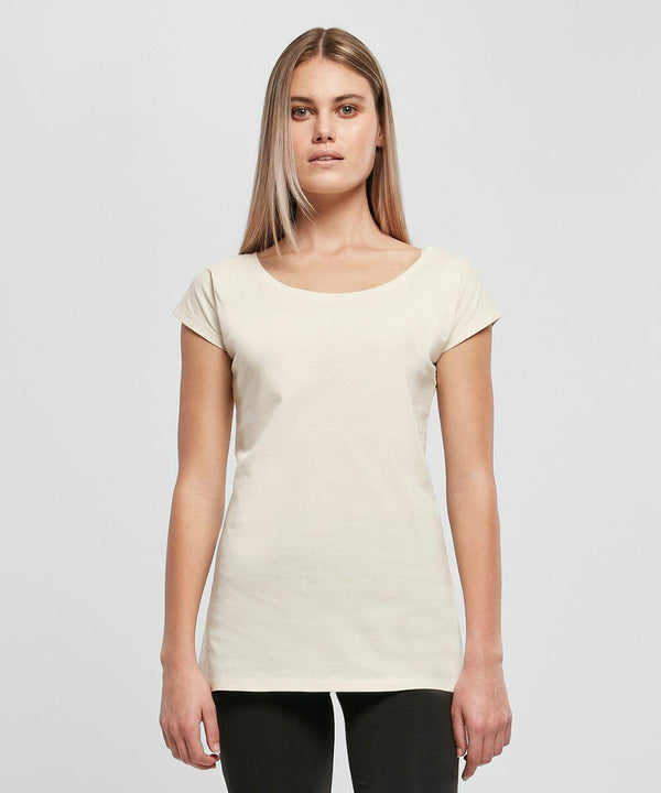 A person with long, straight hair is standing against a plain background. They are wearing a short-sleeve, light-colored BB013 t-shirt from Build Your Brand Basic and dark pants, facing the camera with a neutral expression.
