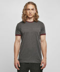 A person with long, curly blond hair stands facing forward against a plain, light gray backdrop. They are wearing a Build Your Brand Basic BB022 gray short-sleeved T-shirt with maroon borders on the sleeves and neckline, paired with dark-colored pants.