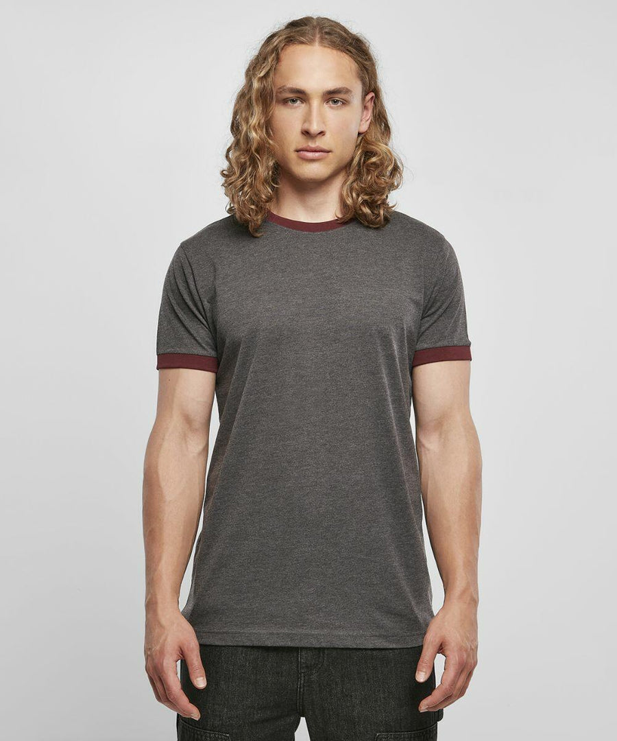 A person with long, curly blond hair stands facing forward against a plain, light gray backdrop. They are wearing a Build Your Brand Basic BB022 gray short-sleeved T-shirt with maroon borders on the sleeves and neckline, paired with dark-colored pants.