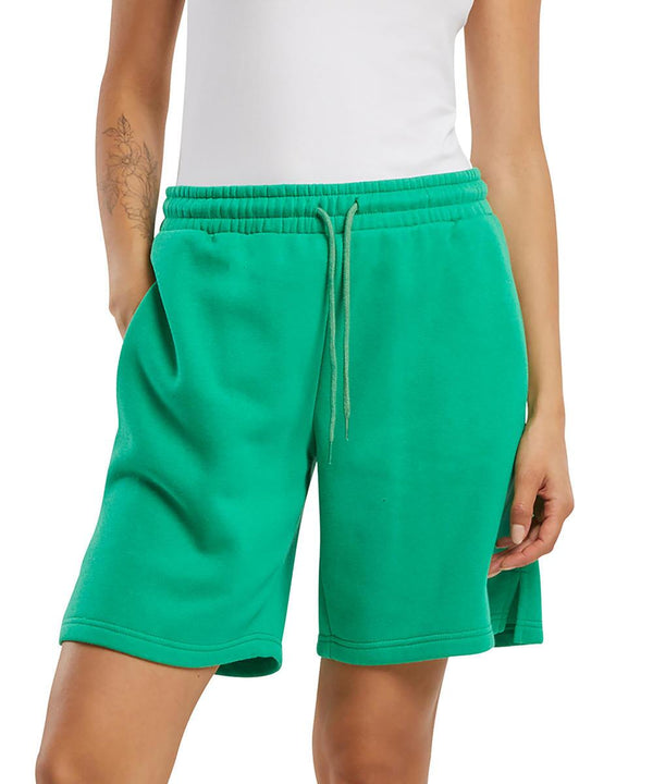 A person wearing Build Your Brand Basic Women's basic sweat shorts in bright green, hands in pockets. Only the lower torso and upper legs are shown, with a floral tattoo visible on the left forearm.