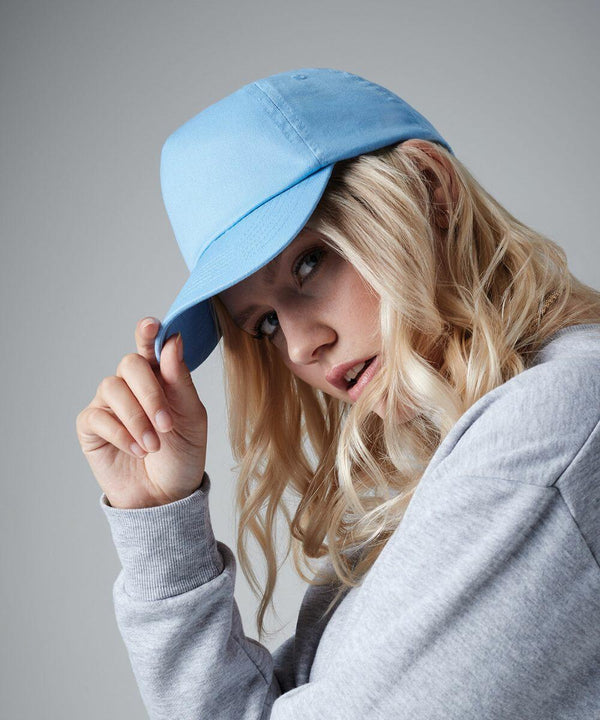 A person with long blonde hair wearing a light blue Beechfield BC010 baseball cap and a gray sweatshirt holds the bill of the cap with their right hand. The person is posed slightly to the side, gazing directly toward the camera against a plain gray background.