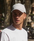 A person wearing a white t-shirt and a light gray Beechfield BC015 baseball cap stands outdoors in a shaded area. The individual has short hair and is sporting gold hoop earrings. The background is blurry, featuring trees and some indistinct structures.