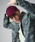 A person wearing a burgundy Beechfield BC025 and camouflage jacket raises one arm as if shielding themselves or adjusting their cap. They have a focused expression and wear a light-colored shirt underneath the jacket. The background is plain gray.