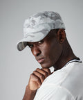 A man wearing a light gray camouflage-patterned Beechfield BC033 cap and a white T-shirt stands against a plain background, with his chin resting on his hand and a contemplative expression on his face.