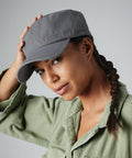 A person wearing a gray Beechfield BC034 cap and a green jacket looks at the camera while slightly tilting the cap with their right hand. They have a long braid over their right shoulder and a calm, neutral expression. The background is plain and gray.