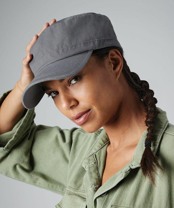 A person wearing a gray Beechfield BC034 cap and a green jacket looks at the camera while slightly tilting the cap with their right hand. They have a long braid over their right shoulder and a calm, neutral expression. The background is plain and gray.