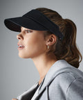 A person with long hair in a ponytail is shown in profile, wearing a black Beechfield BC041 visor and a gray hoodie. They are looking forward with a neutral expression, and small hoop earrings are visible. The background is plain and gray.