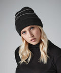 A person with long, wavy blonde hair is wearing a black Beechfield BC042 beanie with white horizontal stripes and a black turtleneck sweater. They have a neutral facial expression and are positioned against a plain gray background.