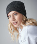 A person with long, wavy blonde hair wears a Beechfield BC044 ribbed dark gray beanie and a white sweater. They are looking into the camera with a slight smile against a plain gray background.
