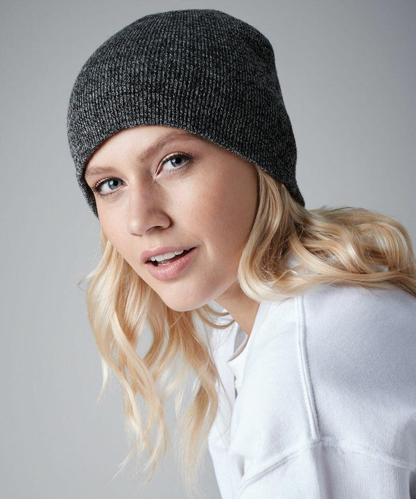 A person with long, wavy blonde hair wears a Beechfield BC044 ribbed dark gray beanie and a white sweater. They are looking into the camera with a slight smile against a plain gray background.