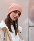 A woman with straight brown hair is wearing a pink Beechfield BC045 beanie and a white jacket. She is looking at the camera with a slight smile. The background is a metallic, reflective surface.