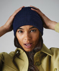 A woman with long hair is wearing a blue knitted Beechfield BC050 and a light green jacket. She has her hands up, adjusting the Beechfield BC050 with a surprised expression. The background is plain and gray.