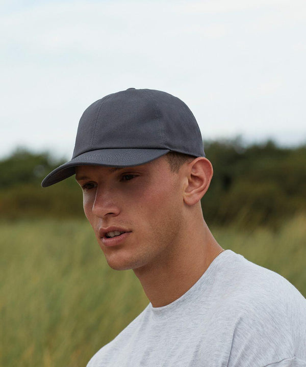 A young man with a neutral expression is wearing a gray Beechfield BC054 and a light gray t-shirt. He is outdoors with a blurred background of greenery and a cloudy sky.