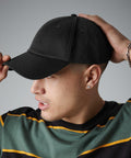 A person with short hair wears a black Beechfield BC059 baseball cap and a green and black striped shirt. Their head is slightly turned to the right, and they have one hand adjusting the cap. They are also wearing a bracelet on one wrist and a stud earring.