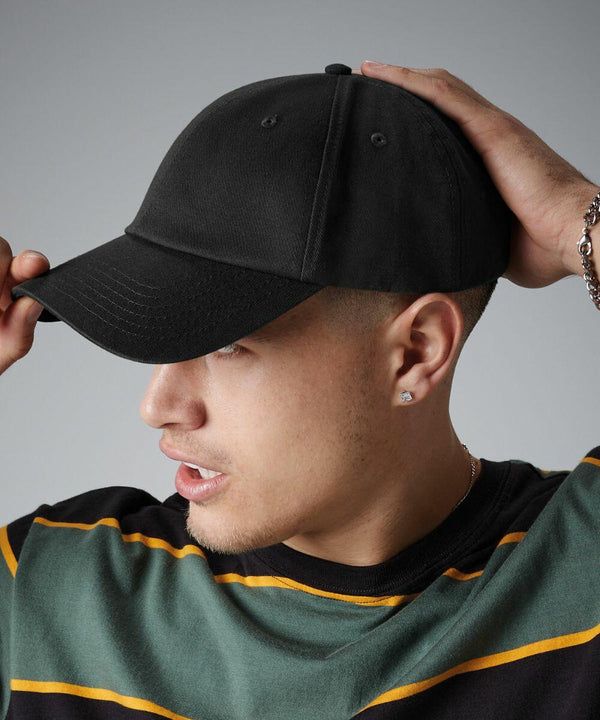 A person with short hair wears a black Beechfield BC059 baseball cap and a green and black striped shirt. Their head is slightly turned to the right, and they have one hand adjusting the cap. They are also wearing a bracelet on one wrist and a stud earring.