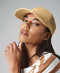 A person with long, dark hair wearing an earthy-toned, striped shirt and a Beechfield BC060 beige cap. They have a thoughtful expression, gazing slightly upward, with their hand resting near their chin. The background is a neutral gray.