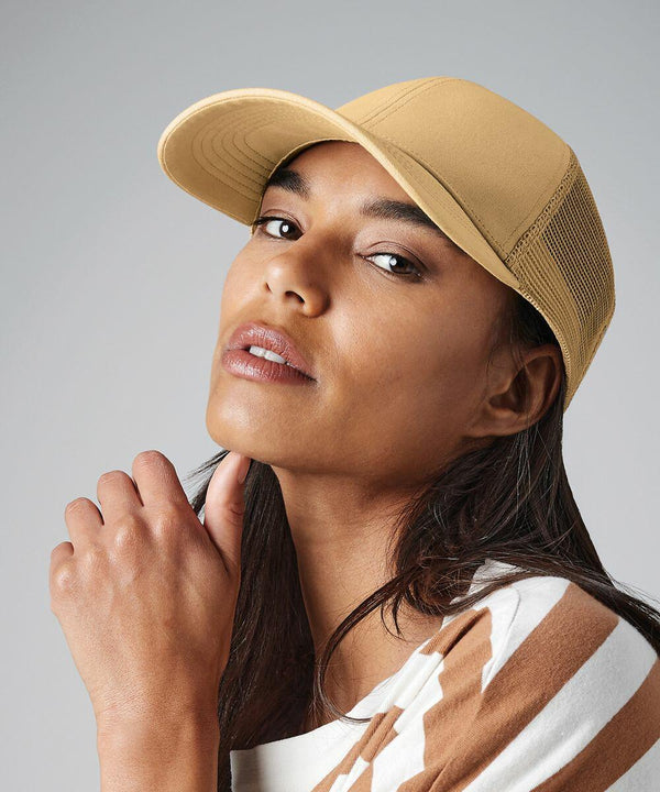 A person with long, dark hair wearing an earthy-toned, striped shirt and a Beechfield BC060 beige cap. They have a thoughtful expression, gazing slightly upward, with their hand resting near their chin. The background is a neutral gray.