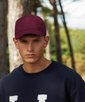 A young man wearing a maroon Beechfield BC065 baseball cap and a dark sweatshirt stands outdoors in a wooded area. He looks directly at the camera with a neutral expression. Tall trees and a bright sky are visible in the background.