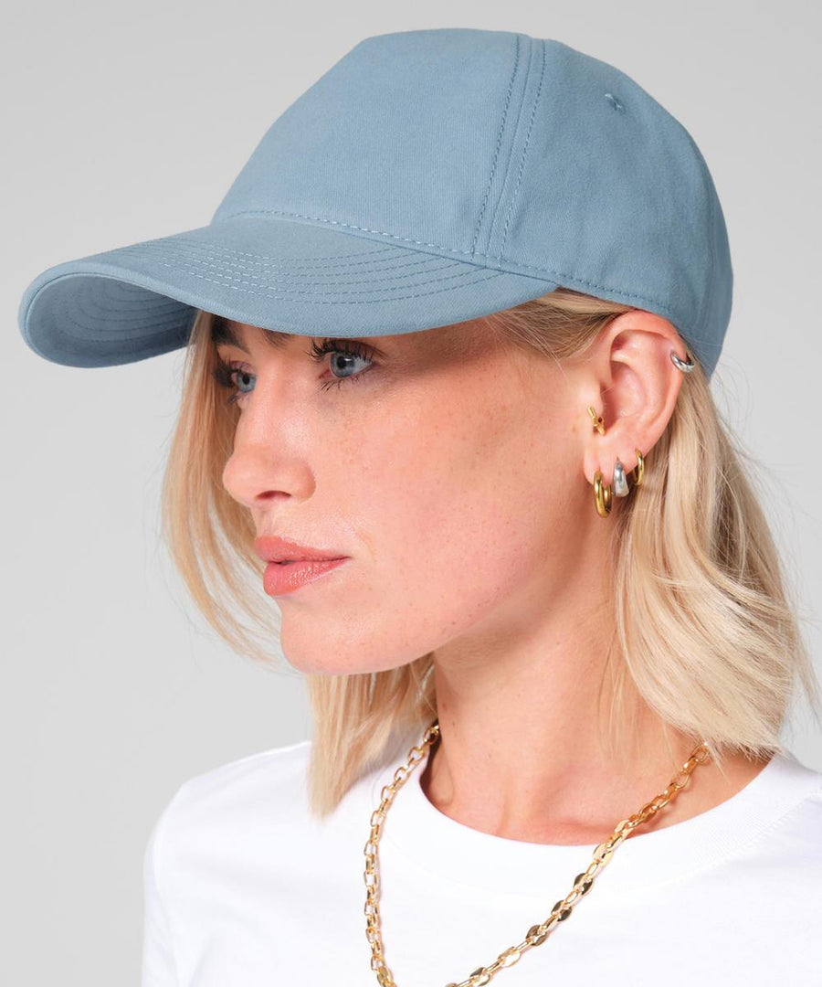 A person with short blonde hair wears a Beechfield Leisure cap in light blue, a white shirt, several gold hoop earrings, and a gold chain necklace, shown in profile against a plain background.