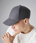 A person with light skin, wearing a Beechfield BC070 gray baseball cap and a white sweater, gazes downward with their hand touching their chin. The background is plain and gray. They have a small earring in their left ear, which is visible in the image.