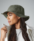 A person with long, dark hair is wearing a green Beechfield BC088 bucket hat and a white jacket over a gray shirt. The individual is looking to the left and holding the collar of the jacket with their right hand. The background is plain and light-colored.