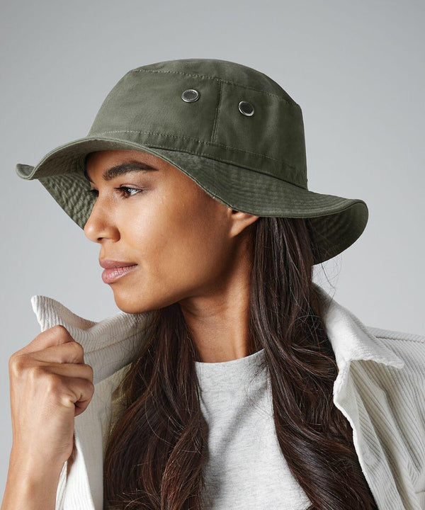 A person with long, dark hair is wearing a green Beechfield BC088 bucket hat and a white jacket over a gray shirt. The individual is looking to the left and holding the collar of the jacket with their right hand. The background is plain and light-colored.