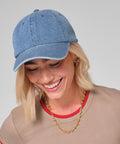 A person with blonde hair wears a Beechfield Denim cap, a tan T-shirt with red trim, a gold chain necklace, and hoop earrings. They are looking down and smiling against a plain light grey background.